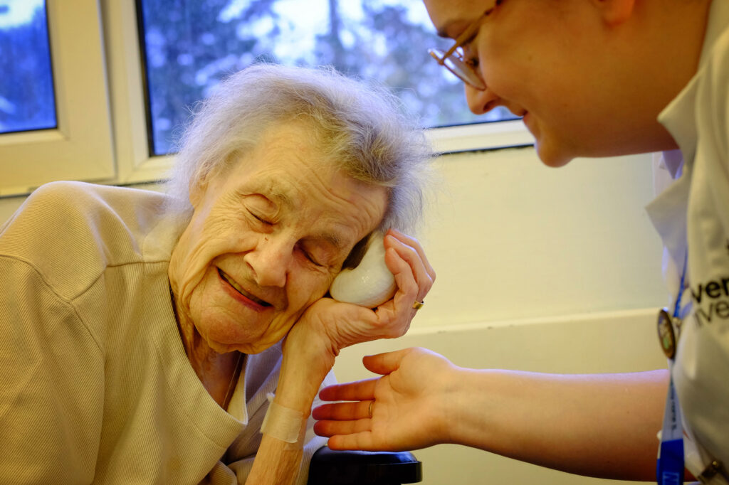 An elderly woman smiles and holds a shell up to her ear. An OT sits beside her smiling and holding up her hand underneath the patient's.