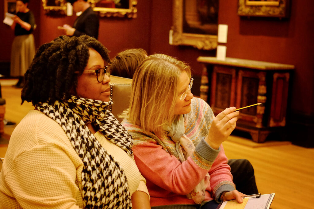 Two women sit on a bench in a red-walled and painting-lined gallery at the Fitzwilliam Museum. One points with a pencil to something in front of her.