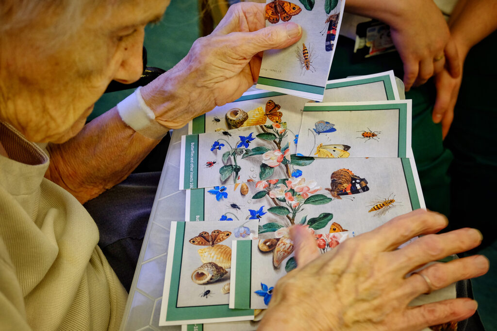 An elderly patient arranges a jigsaw on their lap of a painting of a plant surrounded by butterflies and other insects.