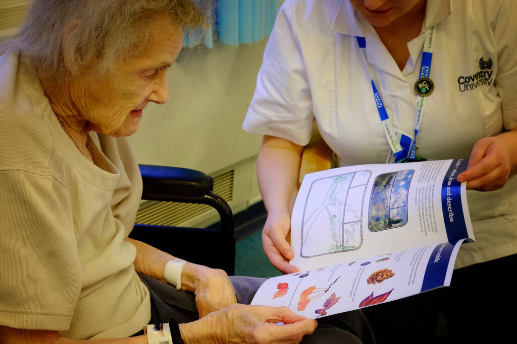An elderly patient in a wheelchair holds an activity sheet held up by an OT sitting next to her.